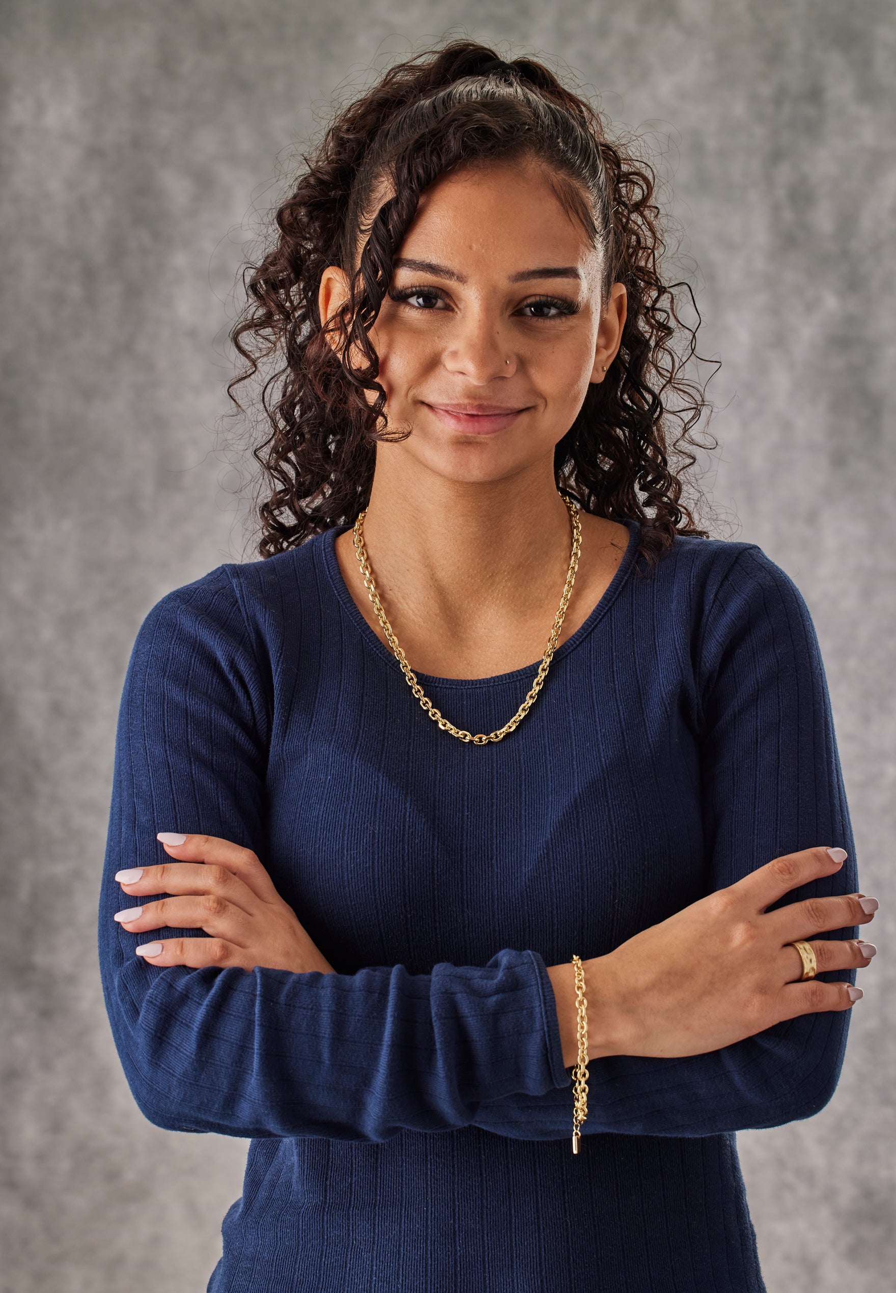 Woman wearing a blue top with gold jewelry against a gray background