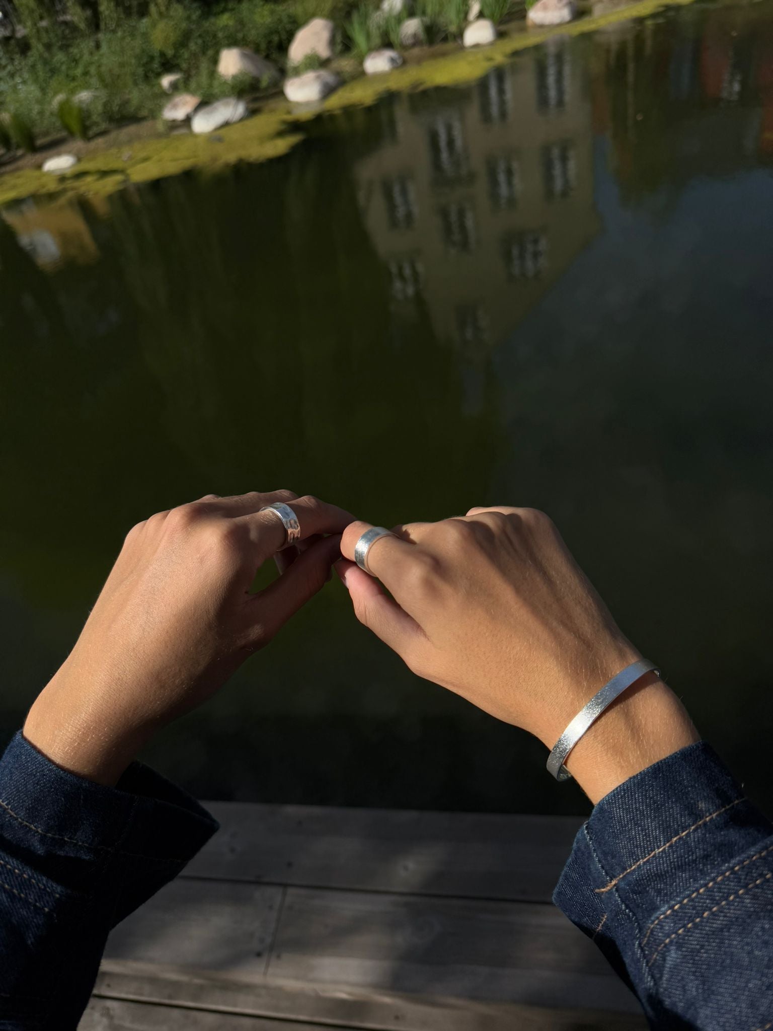 Two hands with rings and a bracelet near a reflective water surface.
