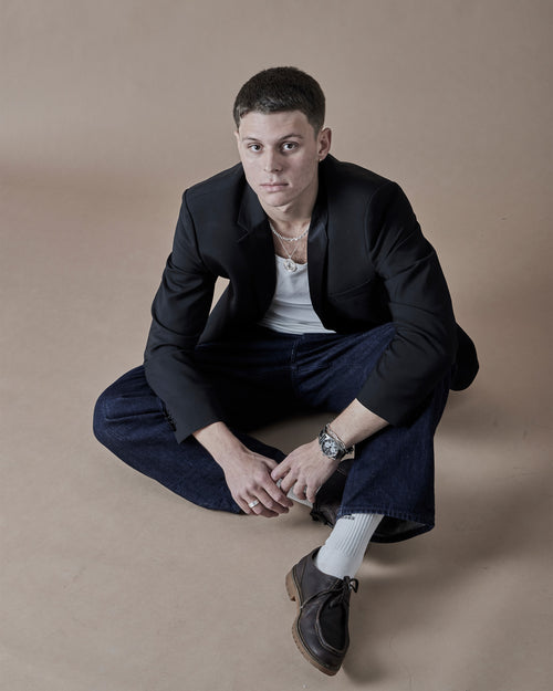 Man sitting cross-legged on a beige background wearing a black jacket and white shirt.
