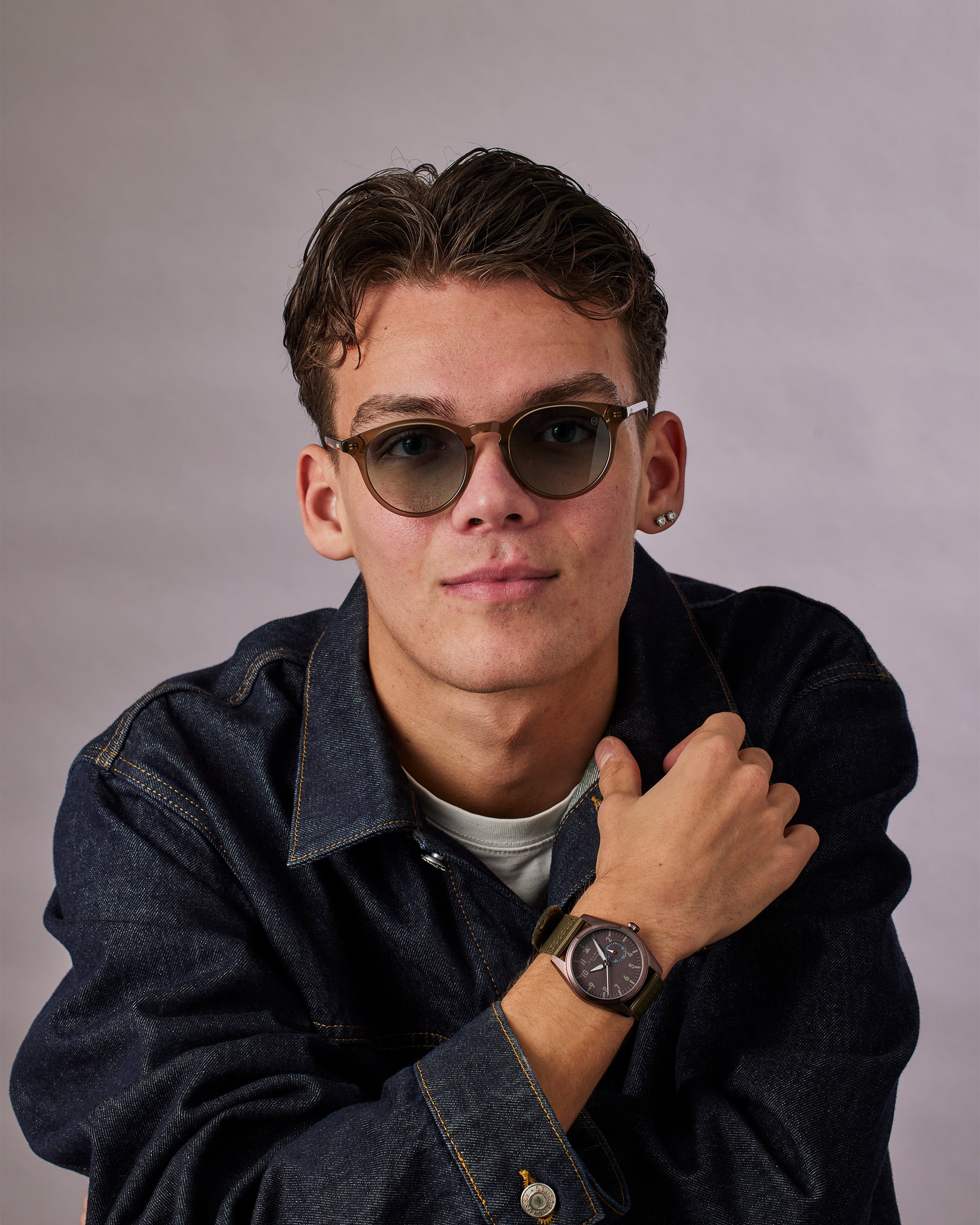 Man wearing sunglasses and a dark jacket with a watch on his wrist against a gray background