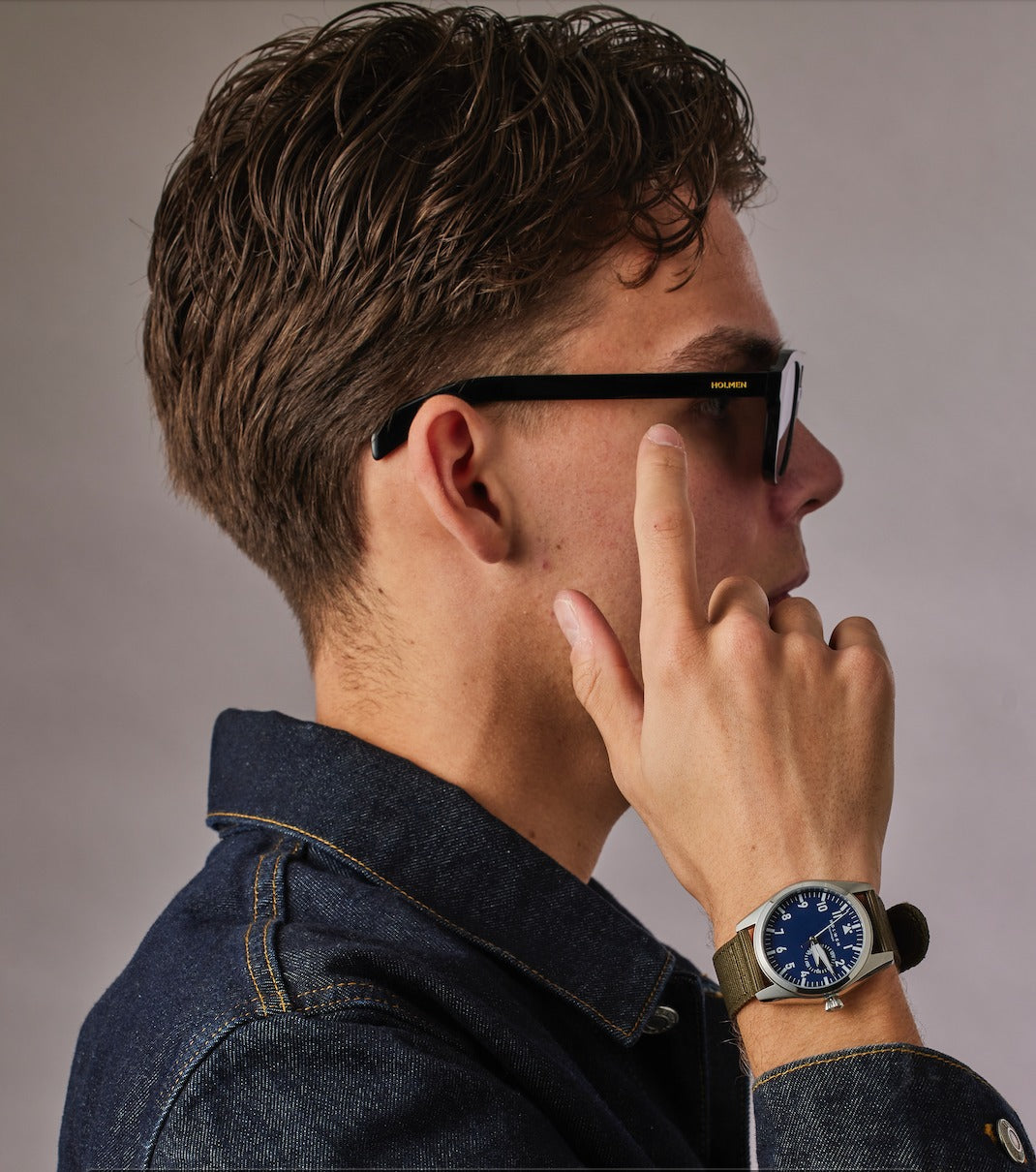 Man wearing a watch with a thoughtful expression, wearing glasses and a denim shirt.
