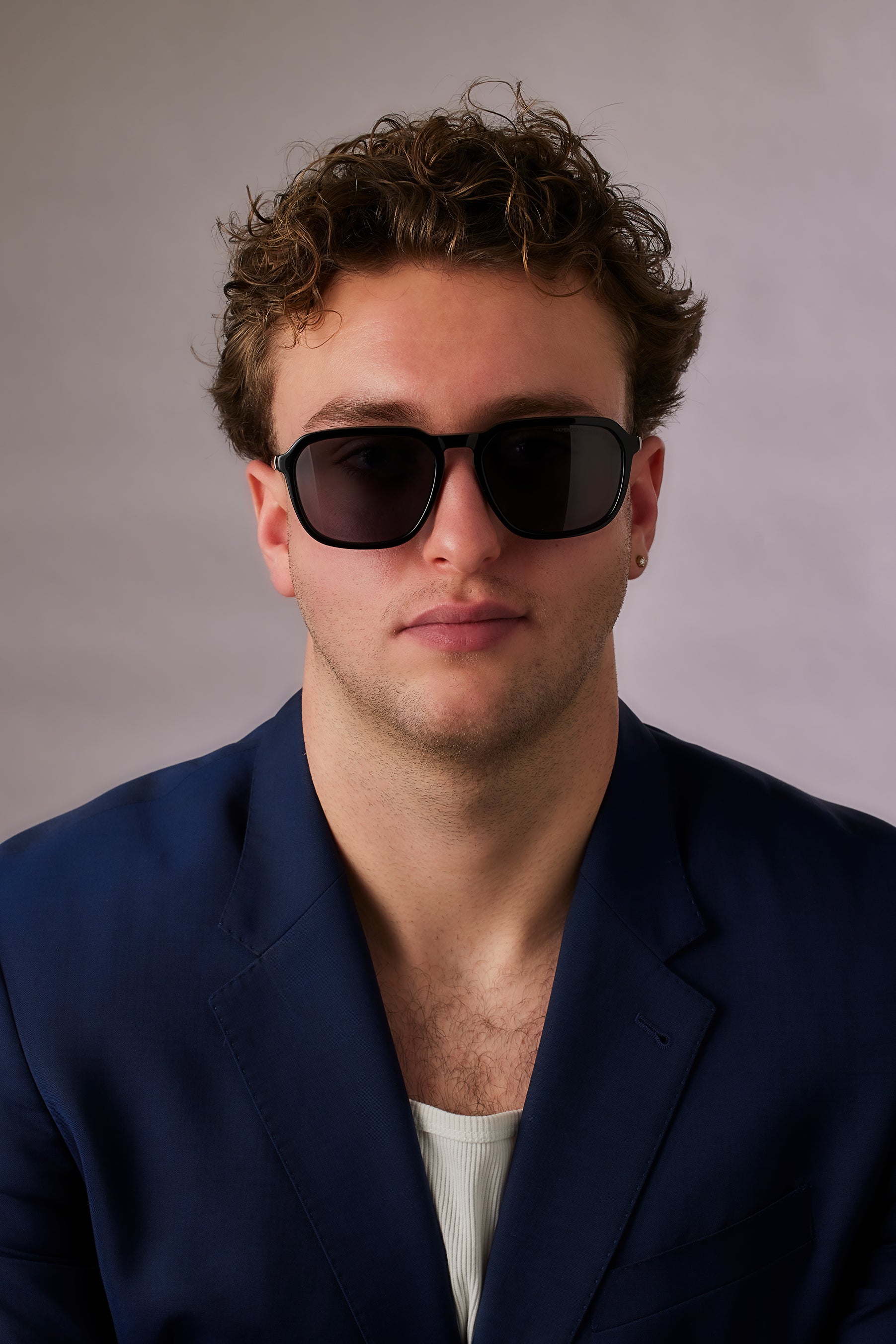 Man wearing sunglasses and a dark blue blazer against a gray background