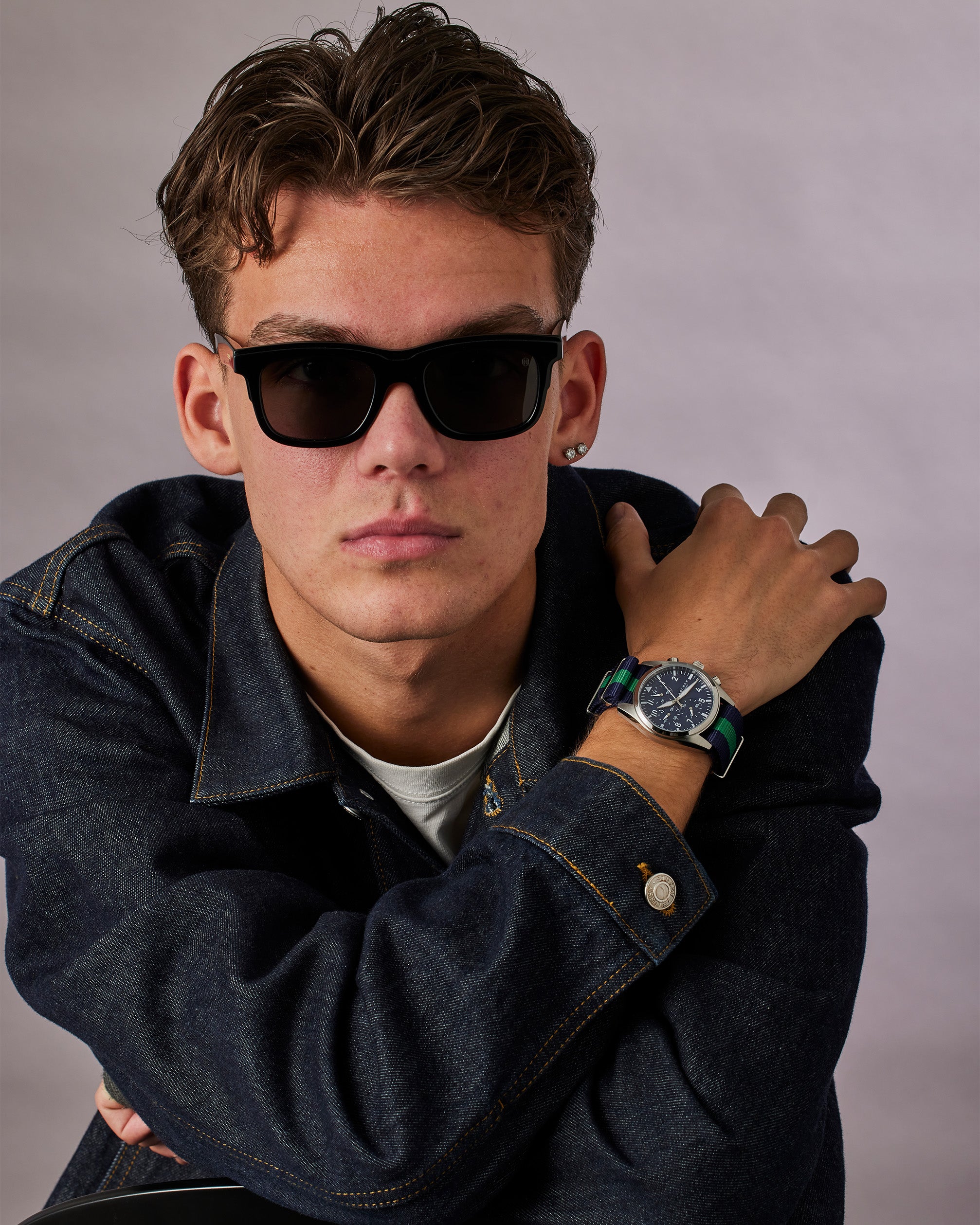 Man wearing sunglasses and a denim jacket with a watch on a gray background